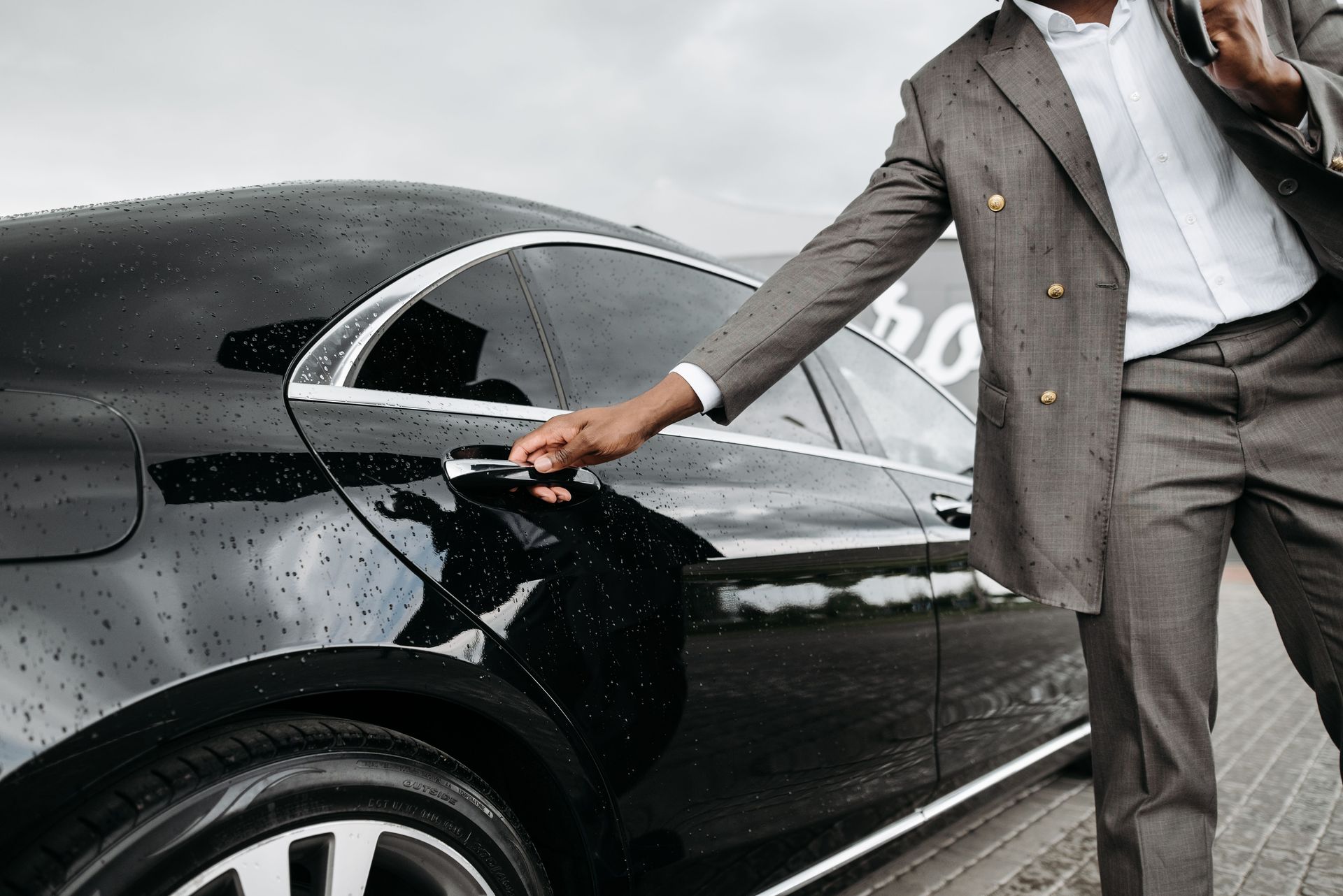 A man in a suit opens a luxury car door under raindrops on stone pavement.